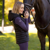 Eine Reiterin in der Seitansicht vor ihrem Pferd, tragend die LeMieux Puffer Gilet Rose in Schwarz. Die leichte Steppweste umschmeichelt die feminine Figur und betont die elegante Quersteppung. Deutlich sichtbar ist die großzügige, gefütterte Kapuze, die zusätzlichen Schutz bietet. Diese schwarze Reitweste wirkt besonders hochwertig und lässt sich universell zu jedem Reitoutfit kombinieren – ein unverzichtbarer Begleiter für die Pferdepflege und die Zeit im Stall.