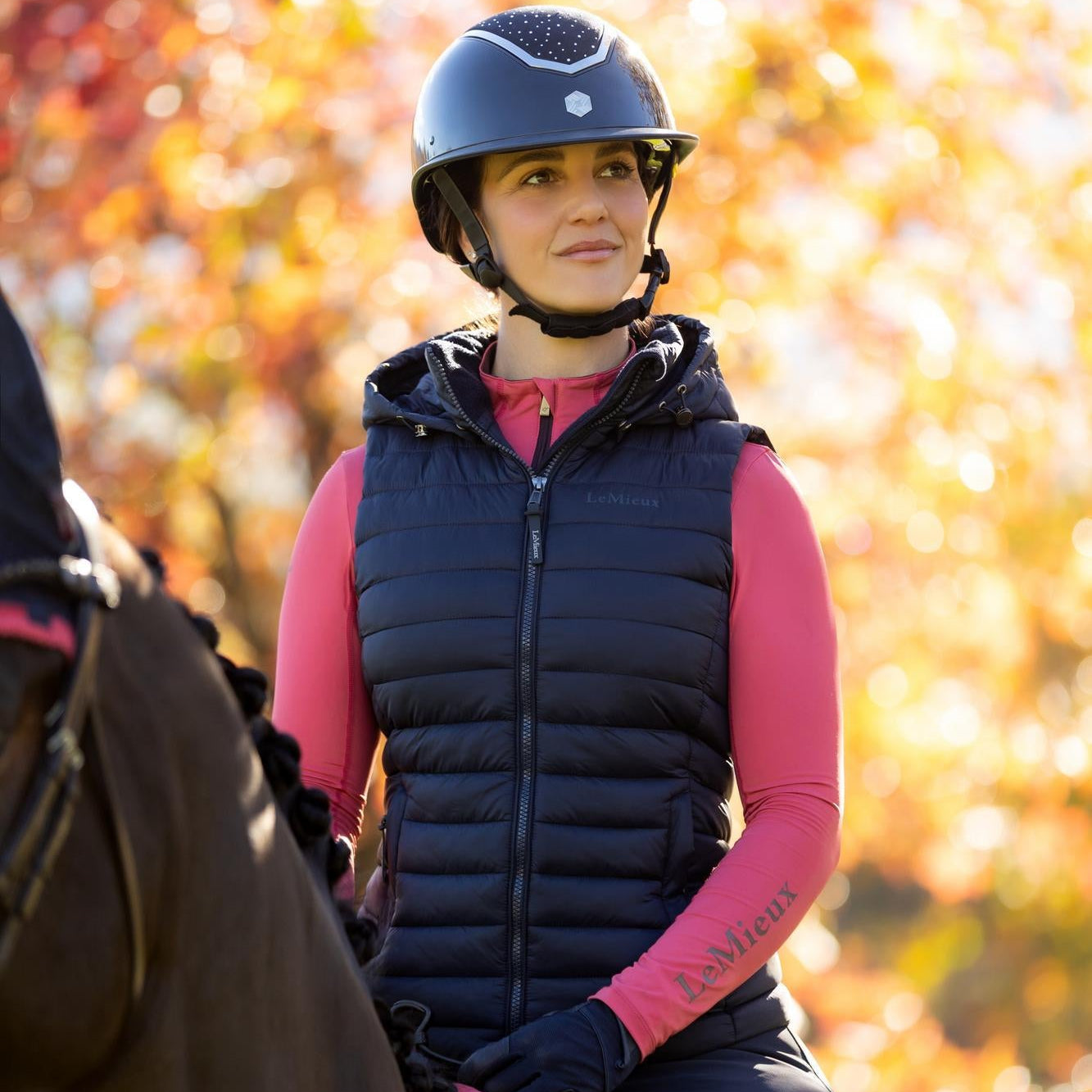 Nahaufnahme der LeMieux Damen Steppweste Rose in Navy während eines herbstlichen Ausritts. Gut sichtbar sind die klassische Quersteppung mit der wärmenden Repreve®-Füllung und die vollständig abnehmbare, verstellbare Kapuze. Der robuste Zwei-Wege-Reißverschluss ist ideal für den Sattel und schützt vor Kratzern. Das elegante Dunkelblau der Pufferweste ist ein vielseitiger Kombinationseinstern für helle oder dunkle Reitoutfits. Funktionale Reitweste mit Marken-Branding bei ePony kaufen.