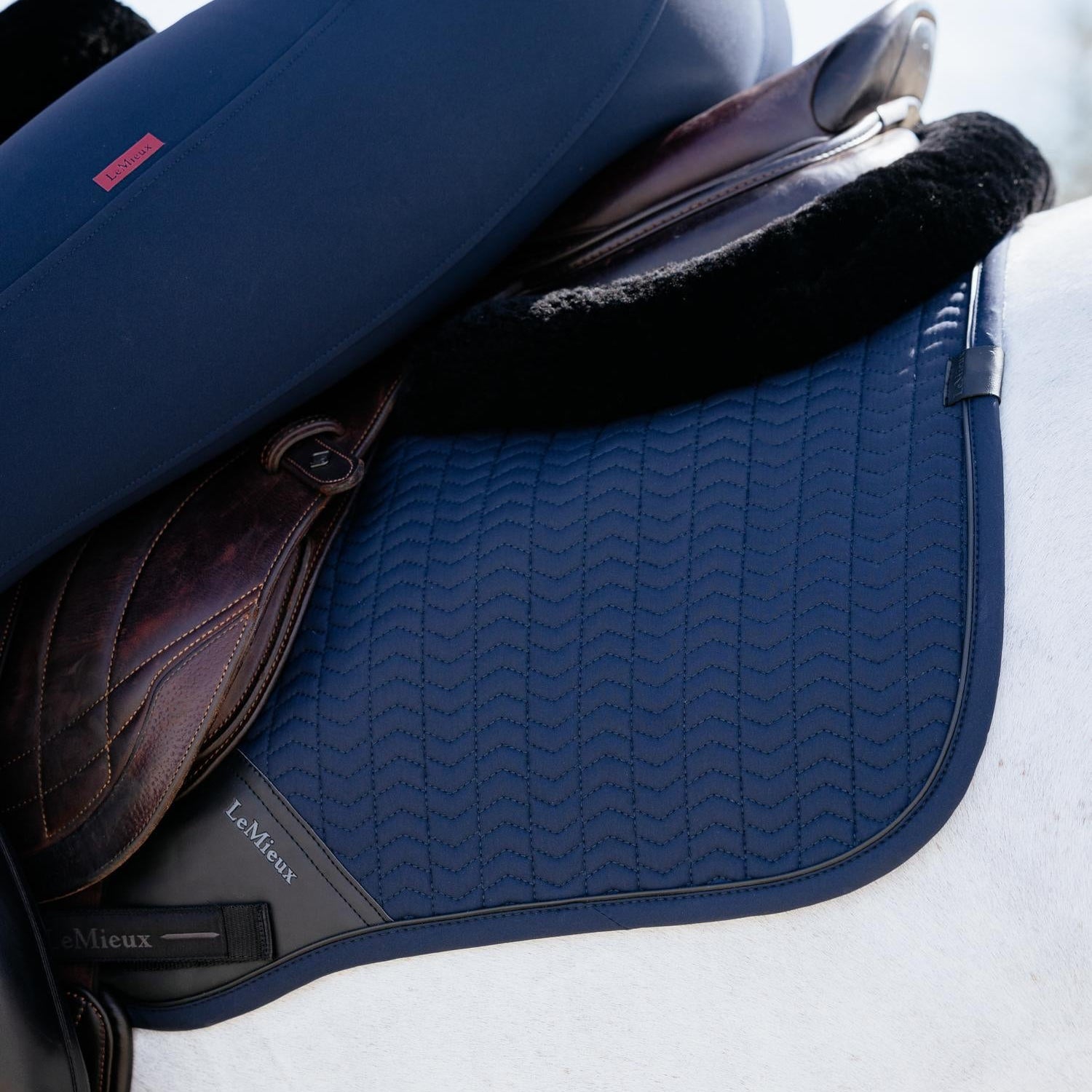 Edler Farbkontrast: Die LeMieux Schabracke Essence Eurojump in Navy liegt zeitlos unter einem schwarzen Springsattel und einem Sattel Half Pad mit Lammfell auf einem Schimmel. Eine dunkle Paspel rahmt die Sattelunterlage in Dunkelblau elegant ein. Das dezente LeMieux Logo ist an der hinteren Logo-Schlaufe zu erkennen und unterstreicht den minimalistischen Look.