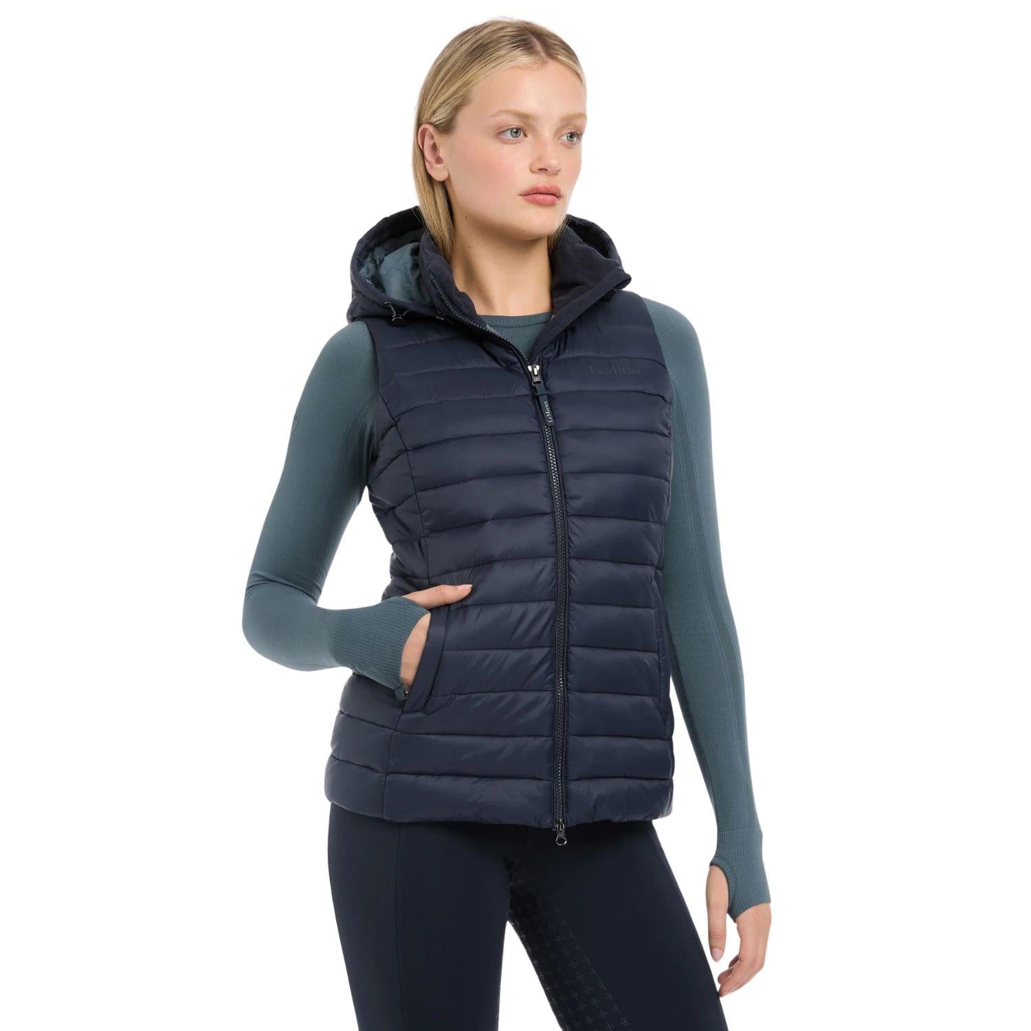 Eine Reiterin trägt die dunkelblaue LeMieux Steppweste Rose in klassischer Quersteppung. Die warme Pufferweste für Damen besticht durch dicke Kammern mit recycelter Füllung und eine moderne Optik. Gut sichtbar ist die abnehmbare, verstellbare Kapuze im Nacken, die Schutz bei Wind bietet. Die Weste umschmeichelt die Silhouette und dient als funktionaler Bodywarmer für kalte Tage im Herbst und Winter bei der Stallarbeit oder beim Training im Sattel. Wasserabweisende Reitkleidung in edlem Navy.
