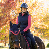 Eine Reiterin beim herbstlichen Ausritt im Wald in der dunkelblauen LeMieux Steppweste Rose mit abnehmbarer Kapuze. Die Damen Reitweste in Navy kombiniert mit einem LeMieux Baselayer zeigt eine optimale Passform im Sattel ohne zu stauchen. Dank der wasserabweisenden Oberfläche und der wärmenden Repreve® Füllung aus recycelten Materialien bleibt der Rumpf bei tief stehender Sonne perfekt isoliert. Hochwertige Pufferweste mit Quersteppung für Training und Freizeit im Pferdesport bei ePony kaufen.