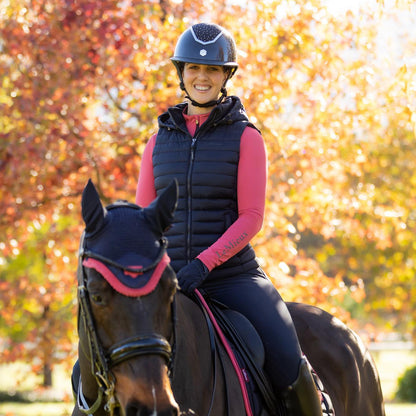 Eine Reiterin beim herbstlichen Ausritt im Wald in der dunkelblauen LeMieux Steppweste Rose mit abnehmbarer Kapuze. Die Damen Reitweste in Navy kombiniert mit einem LeMieux Baselayer zeigt eine optimale Passform im Sattel ohne zu stauchen. Dank der wasserabweisenden Oberfläche und der wärmenden Repreve® Füllung aus recycelten Materialien bleibt der Rumpf bei tief stehender Sonne perfekt isoliert. Hochwertige Pufferweste mit Quersteppung für Training und Freizeit im Pferdesport bei ePony kaufen.