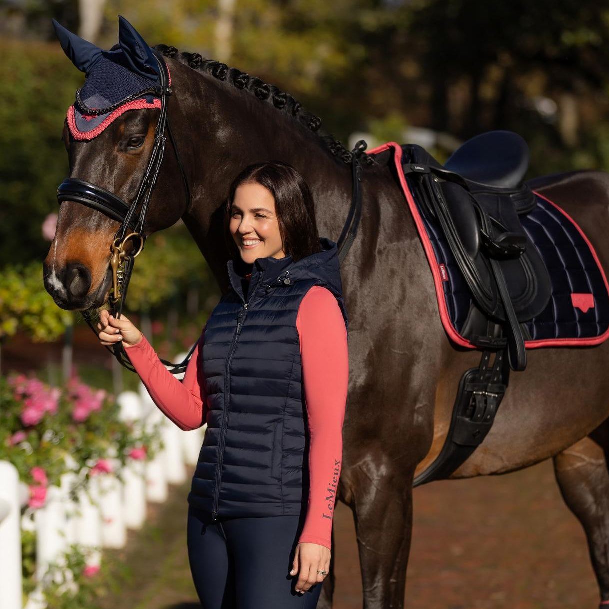 Eine Reiterin steht elegant neben ihrem Pferd in einem perfekt abgestimmten LeMieux Total Look in schimmerndem Navy. Die LeMieux Damen Steppweste Rose mit ihrer feinen Quersteppung und wärmender Repreve®-Füllung schimmert edel im Sonnenlicht. Das Outfit wird komplettiert durch eine farblich passende LeMieux Schabracke und Fliegenmütze in Dunkelblau, die ebenfalls einen luxuriösen Glanz aufweisen. Diese harmonische Kombination definiert Reitmode neu und bietet höchsten Komfort und Stil für Reiterinnen.