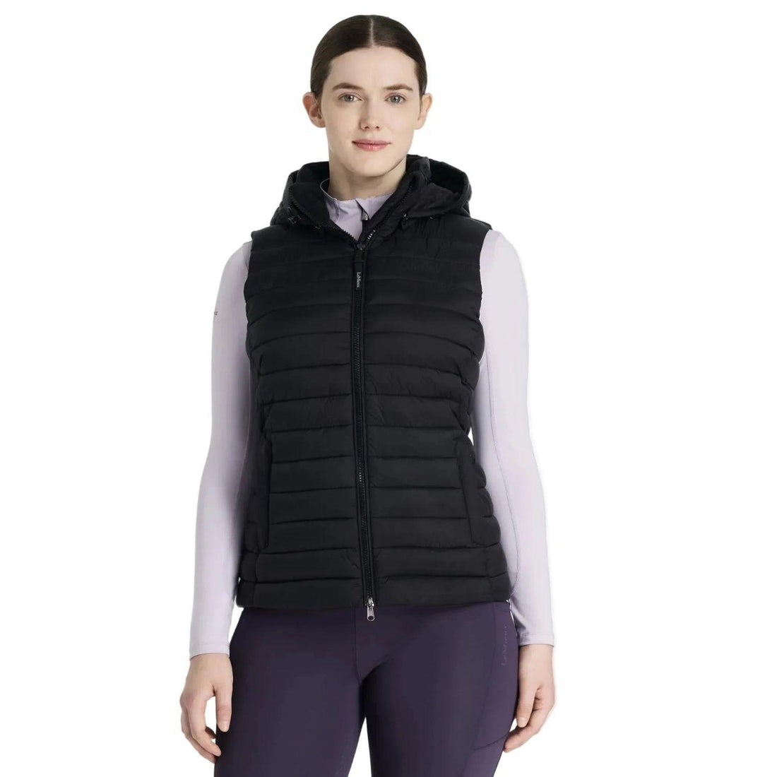 Eine Reiterin trägt die schwarze LeMieux Steppweste Rose in klassischer Quersteppung. Die warme Pufferweste für Damen besticht durch dicke Kammern mit recycelter Füllung und eine moderne Optik. Gut sichtbar ist die abnehmbare, verstellbare Kapuze im Nacken, die Schutz bei Wind bietet. Die Weste umschmeichelt die Silhouette und dient als funktionaler Bodywarmer für kalte Tage im Herbst und Winter bei der Stallarbeit oder beim Training im Sattel. Wasserabweisende Reitkleidung in edlem Schwarz.