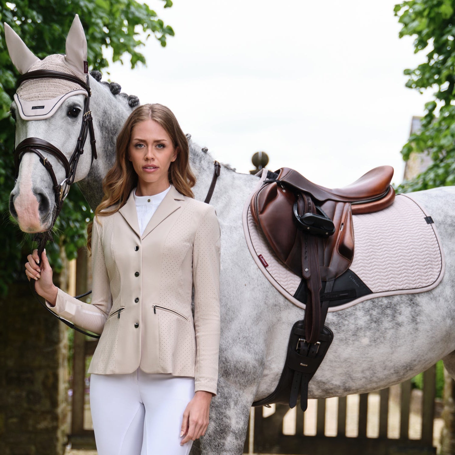 Perfektes Match für das Springturnier an heißen Sommertagen: Eine Reiterin im atmungsaktiven LeMieux Turnierjacket Jessica Mesh in Stone steht neben ihrem Pferd mit der passende LeMieux Essence Eurojump Schabracke in Stone. Das harmonische Outfit in edlem Beige zeigt, wie elegant die Farbe Stone das klassische Weiß als Turnierfarbe ersetzt und gleichzeitig höchsten Komfort durch Mesh-Material bietet.