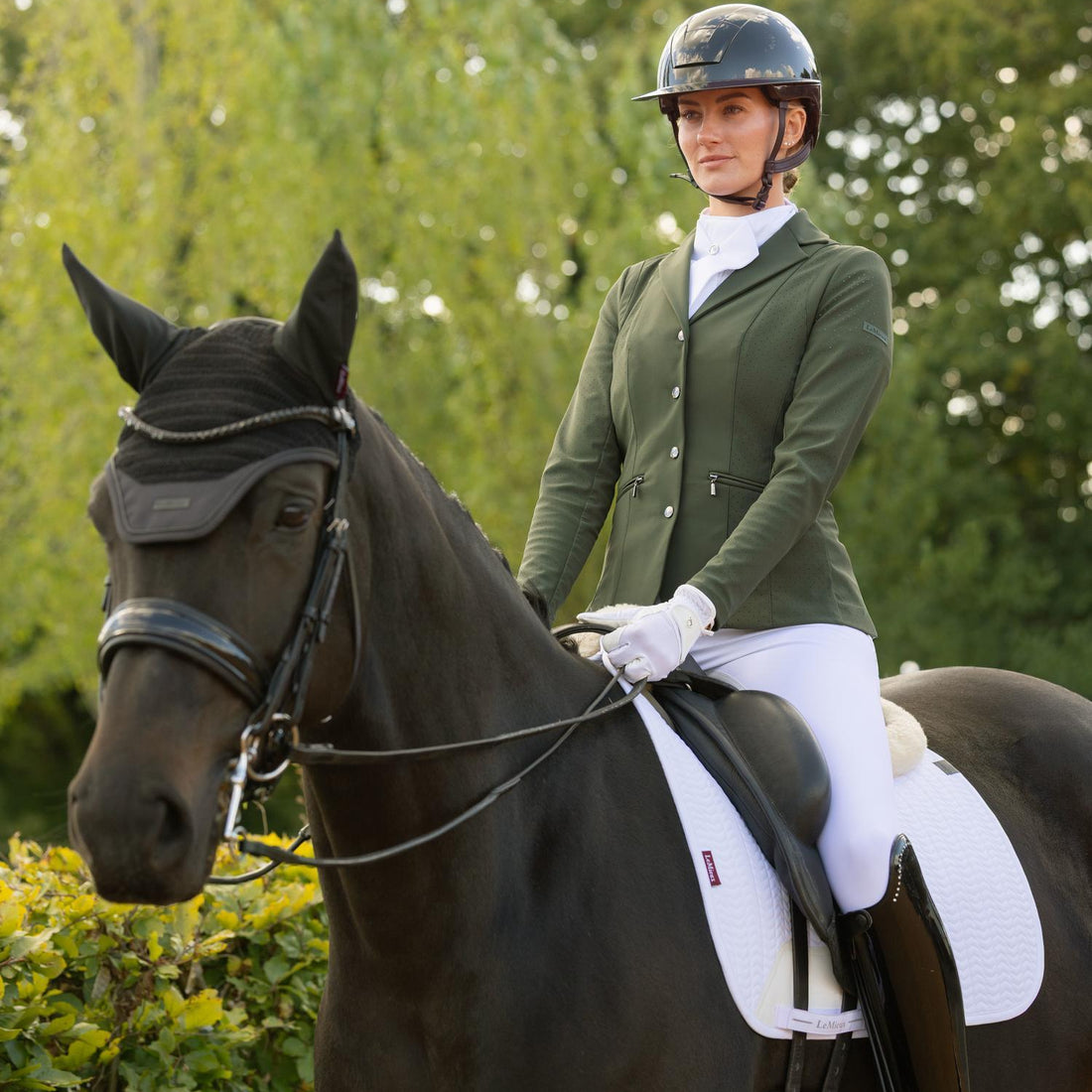 Turnier-Ready: Eine Reiterin in voller Turnierkleidung sitzt auf einem gesattelten Dressurpferd mit Kandaren-Zäumung. Die LeMieux Essence Dressurschabracke in strahlendem Weiß liegt perfekt unter dem Dressursattel und passt sich dem Pferderücken an. Die Szenerie strahlt höchste Eleganz kurz vor dem Einreiten in die Dressurprüfung aus.