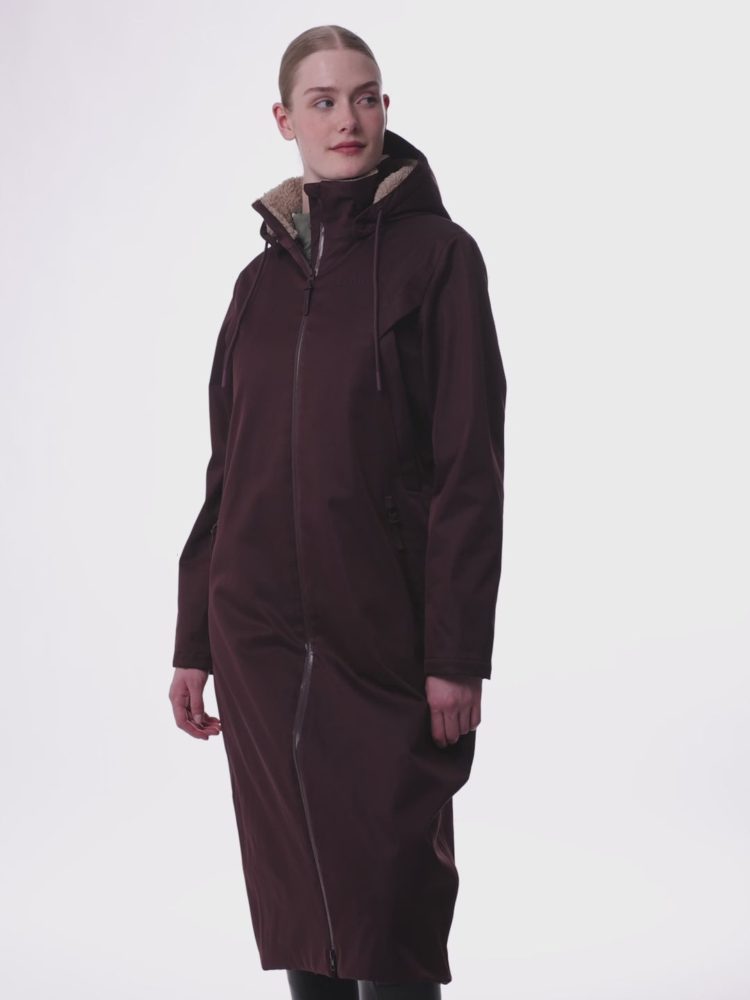 Junge blonde Frau trägt den LeMieux Reitmantel All Elements in der Farbe damson geschlossen und zeigt diesen von vorne, von der Seite und hinten. Am Kragen und der Kapuze ist das beigefarbene Teddy Fleece zu sehen. Der weite lockere Schnitt ist gut zu sehen und auch der Rückseitige Schlitz  der zum Reiten geöffnet werden kann. Das LeMieux Logo auf der linken Brust ist sehr dezent. Die Taschen sind durch Reißverschlüsse alle geschlossen. Der Reitmantel wirkt weit, locker und am Kragen weich und plüschig.