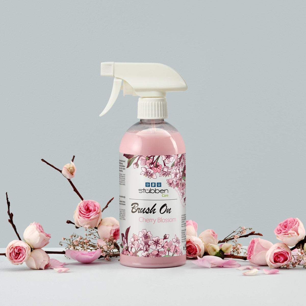 Stübben Brush On Cherry Blossom 500 ml Sprühflasche im Stillleben mit Kirschblütenzweigen und Rosenblüten. Mähnenspray und Schweifspray mit Kirschblüten-Design für Glanz, Volumen und leichte Kämmbarkeit.