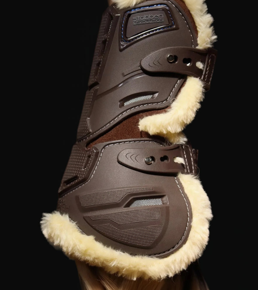Nahaufnahme der braunen Stübben Evolution Hybrid Gamasche von der Seite: Die stoßabsorbierende TPE-Hartschale schützt die Beugesehne zuverlässig vor Tritten. Gut erkennbar ist die ergonomische Aussparung für maximale Fesselgelenks-Freiheit sowie die vertikalen Belüftungskanäle zur Thermoregulation. Die elastischen Schnellverschlüsse mit stabilen Pin-Knöpfen garantieren eine individuelle Passform und verhindern störende Druckpunkte am Pferdebein während der Springgymnastik.