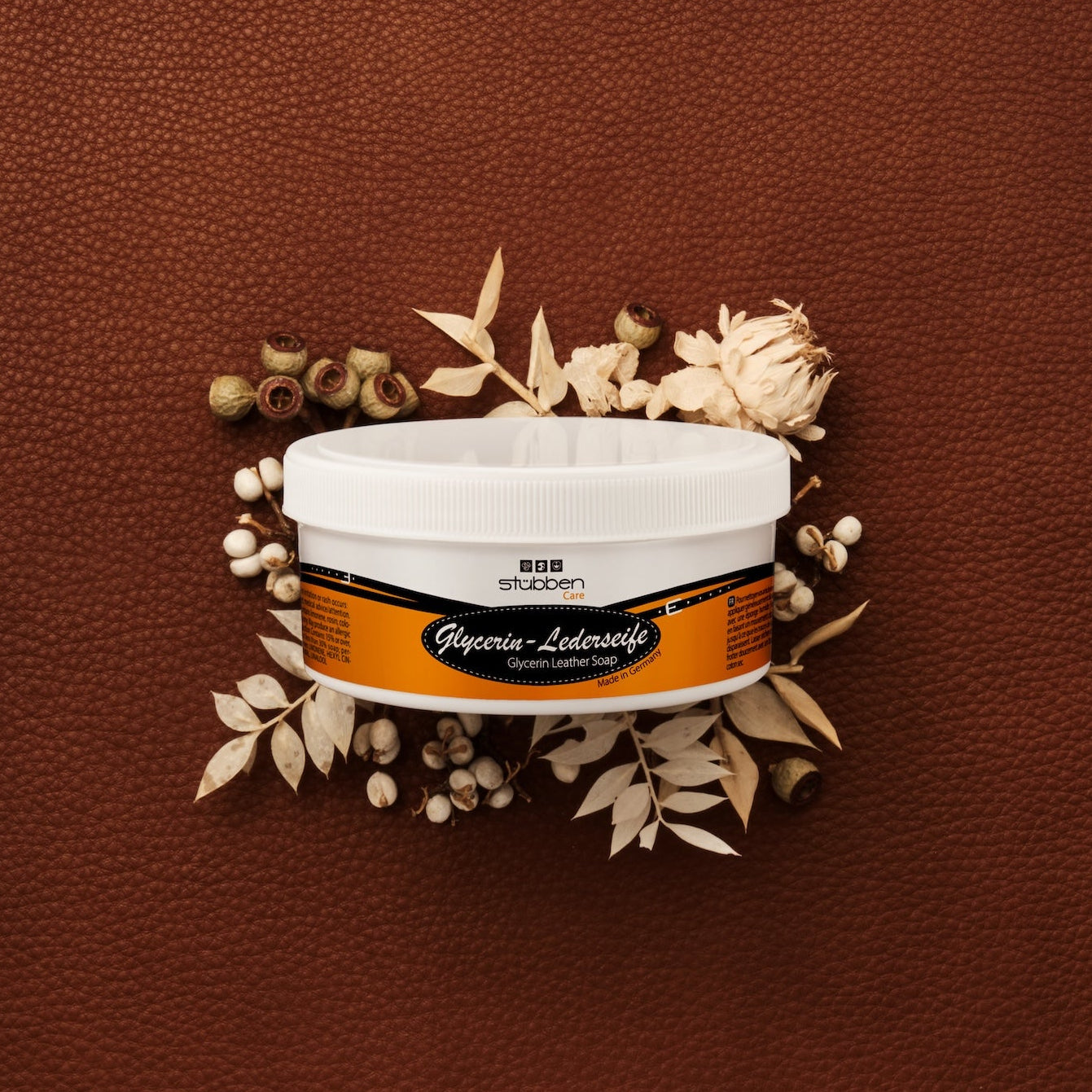 Die 250 g Dose Stübben Glycerin Sattelseife steht vor einem rustikalen, groben braunen Lederhintergrund, der durch helle Trockenblumen kontrastiert wird, um die sanfte Reinigung ohne Austrocknung und den Erhalt der Lederelastizität zu symbolisieren.