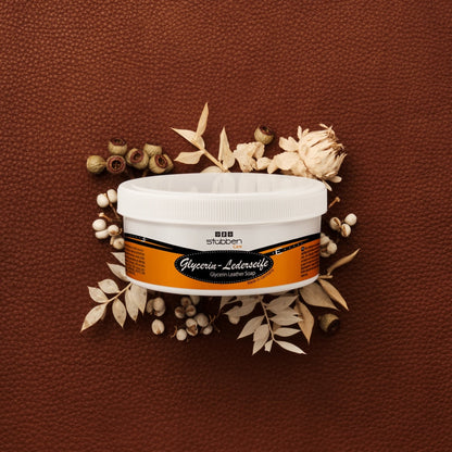 Die 250 g Dose Stübben Glycerin Sattelseife steht vor einem rustikalen, groben braunen Lederhintergrund, der durch helle Trockenblumen kontrastiert wird, um die sanfte Reinigung ohne Austrocknung und den Erhalt der Lederelastizität zu symbolisieren.