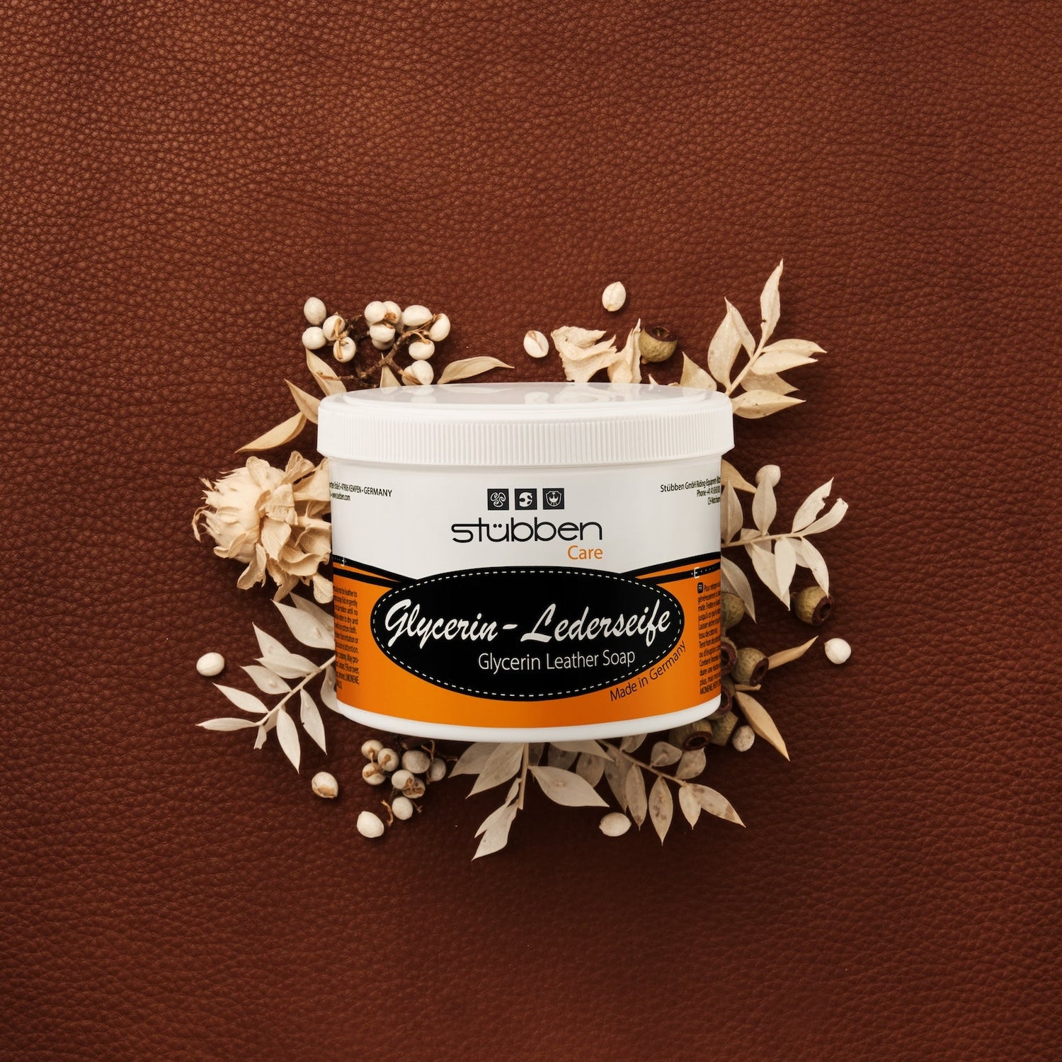 Vor einem groben braunen Lederhintergrund mit hellen Trockenblumen-Akzenten präsentiert sich die ergiebige 500 g Vorratsdose der Stübben Glycerin Sattelseife, die für geschmeidiges Leder und langanhaltende Elastizität durch rückfettende Reinigung steht.