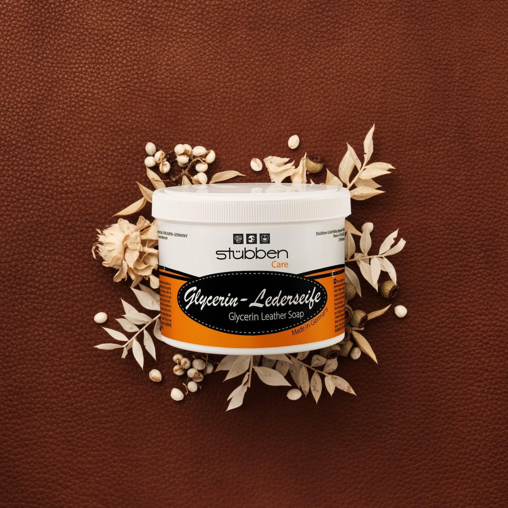 Vor einem groben braunen Lederhintergrund mit hellen Trockenblumen-Akzenten präsentiert sich die ergiebige 500 g Vorratsdose der Stübben Glycerin Sattelseife, die für geschmeidiges Leder und langanhaltende Elastizität durch rückfettende Reinigung steht.