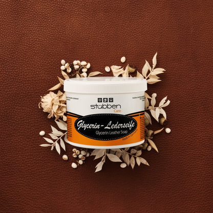 Vor einem groben braunen Lederhintergrund mit hellen Trockenblumen-Akzenten präsentiert sich die ergiebige 500 g Vorratsdose der Stübben Glycerin Sattelseife, die für geschmeidiges Leder und langanhaltende Elastizität durch rückfettende Reinigung steht.