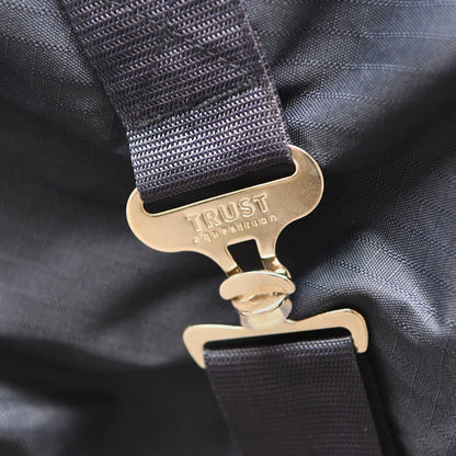 Diese Detailaufnahme zeigt die stabilen, metallischen T-Verschlüsse der gekreuzten Bauchgurte mit der exklusiven Trust Equestrian Aufschrift, die für eine sichere Fixierung der 100g Outdoordecke sorgen und durch ihre robuste Verarbeitung eine langlebige Positionierung des Tetris-Systems am Pferdekörper garantieren.
