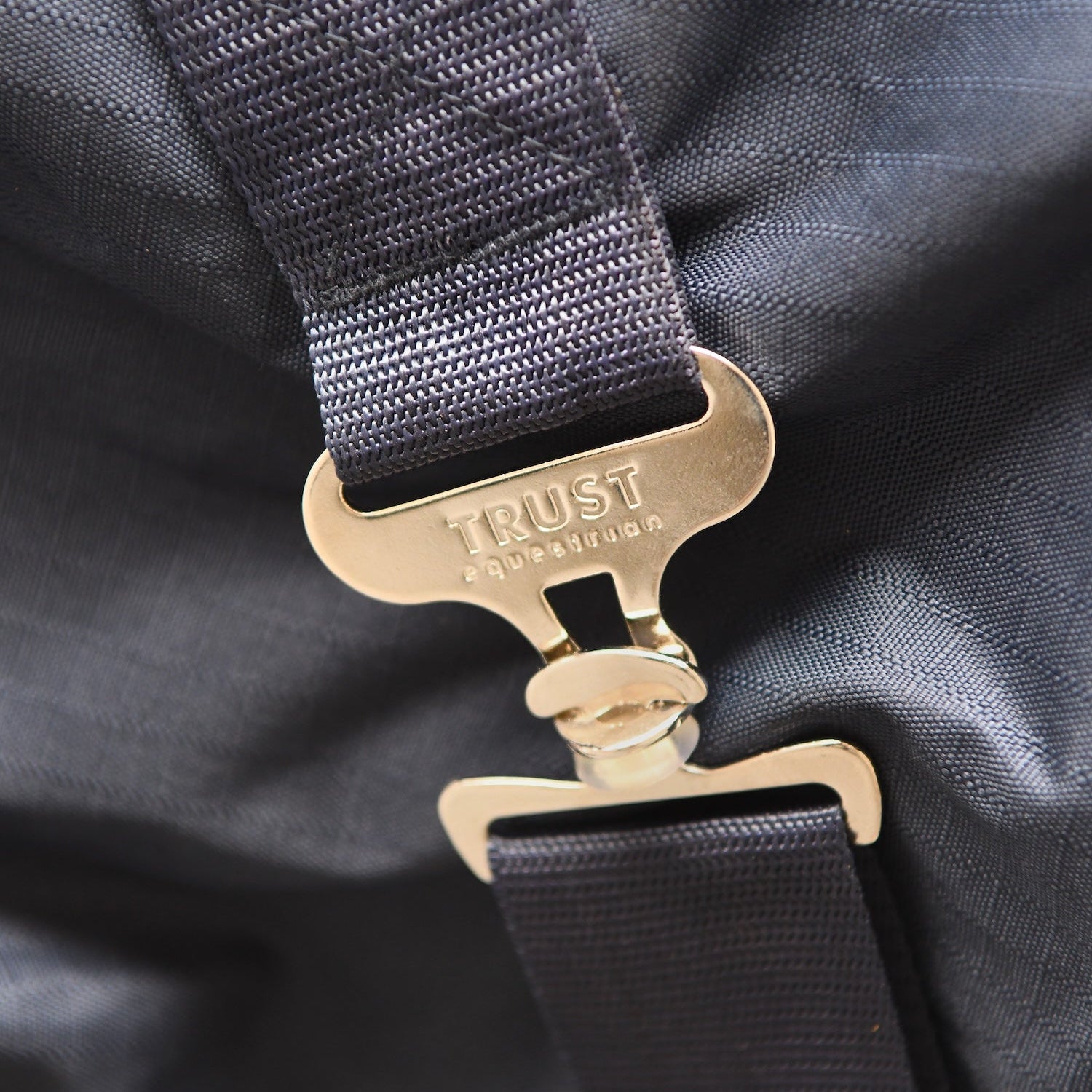Diese Detailaufnahme zeigt die stabilen, metallischen T-Verschlüsse der gekreuzten Bauchgurte mit der exklusiven Trust Equestrian Aufschrift, die für eine sichere Fixierung der 300g Outdoordecke sorgen und durch ihre robuste Verarbeitung eine langlebige Positionierung des Tetris-Systems am Pferdekörper garantieren.