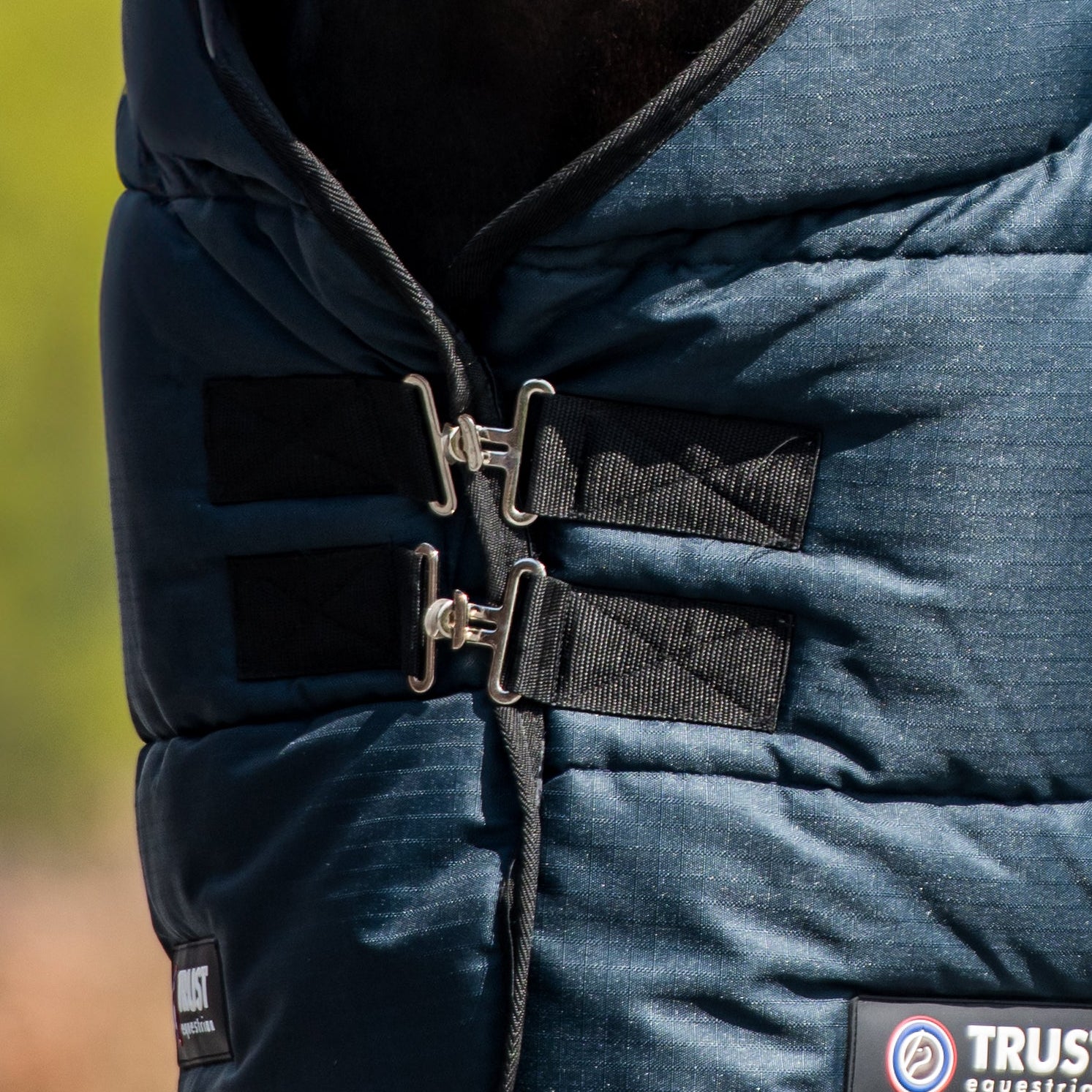 Die Detailaufnahme zeigt den stabilen Brustverschluss der Trust Equestrian Tetris Stalldeckedecke mit zwei robusten, metallischen T-Verschlüssen plus Sicherheitsgummis inklusive Trust-Logo-Prägung, die für einen sicheren Halt in der Brustposition sorgen und ein Verrutschen der 200g Boxendecke verhindern.