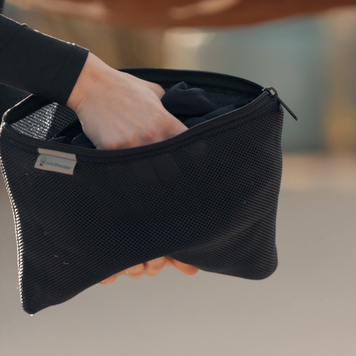 Nahaufnahme der originalen Waldhausen Netztasche mit Markenlabel: Eine Hand greift in die geöffnete schwarze Reißverschlusstasche, um die vollgesaugte Waldhausen Health + Care Ice-Layer Kühldecke zu entnehmen. Nach dem Tränken im Eimer wird die Decke herausgezogen, ausgewrungen und zur effektiven Verdunstungskühlung direkt auf den Pferderücken gelegt. Handlich zur Anwendung unterwegs, auch bei Turnieren.