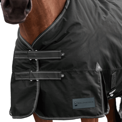 Detailaufnahme des doppelten Brustverschlusses der Waldhausen Outdoordecke Arctic 50 g in Schwarz. Die robusten T-Verschlüsse sind mit Klettabdeckungen geschützt, und können ein Hängenbleiben im Offenstall oder Paddock reduzieren. Der doppelte Brustverschluss ermöglicht einfaches Ein- und Ausdecken von Pferd oder Pony. Reflektierende Kontrastnähte in Weiß auf den Klettlaschen und dem Einfassband erhöhen die Sichtbarkeit bei Dunkelheit.