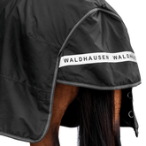 Rückansicht der dunklen Waldhausen-Outdoordecke Arctic 100 g in Schwarz mit Fokus auf den langen Schweiflatz. Der großzügige Schweiflatz schützt Schweif und Schweifrübe des Pferdes zuverlässig vor Wind, Regen und Kälte. Die breite, reflektierende Waldhausen-Applikation sorgt für optimale Sichtbarkeit von hinten im Dunkeln, auf der Weide, auf Wegen oder im Straßenverkehr. Die perfekte 600D-Ripstop-Regendecke für bessere Sichtbarkeit im Dunklen. D-Ringe für Beinschnüre sind zu sehen.
