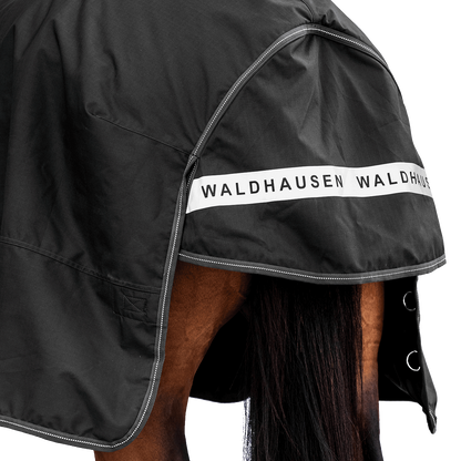Rückansicht der dunklen Waldhausen-Outdoordecke Arctic 100 g in Schwarz mit Fokus auf den langen Schweiflatz. Der großzügige Schweiflatz schützt Schweif und Schweifrübe des Pferdes zuverlässig vor Wind, Regen und Kälte. Die breite, reflektierende Waldhausen-Applikation sorgt für optimale Sichtbarkeit von hinten im Dunkeln, auf der Weide, auf Wegen oder im Straßenverkehr. Die perfekte 600D-Ripstop-Regendecke für bessere Sichtbarkeit im Dunklen. D-Ringe für Beinschnüre sind zu sehen.