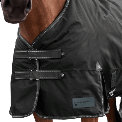 Detailaufnahme des doppelten Brustverschlusses der Waldhausen Outdoordecke Arctic 200 g in Schwarz. Die robusten T-Verschlüsse sind mit Klettabdeckungen geschützt, und können ein Hängenbleiben im Offenstall oder Paddock reduzieren. Der doppelte Brustverschluss ermöglicht einfaches Ein- und Ausdecken von Pferd oder Pony. Reflektierende Kontrastnähte in Weiß auf den Klettlaschen und dem Einfassband erhöhen die Sichtbarkeit bei Dunkelheit.