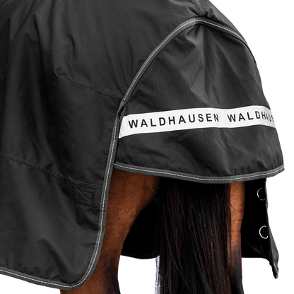 Rückansicht der dunklen Waldhausen-Outdoordecke Arctic 200 g in Schwarz mit Fokus auf den langen Schweiflatz. Der großzügige Schweiflatz schützt Schweif und Schweifrübe des Pferdes zuverlässig vor Wind, Regen und Kälte. Die breite, reflektierende Waldhausen-Applikation sorgt für optimale Sichtbarkeit von hinten im Dunkeln, auf der Weide, auf Wegen oder im Straßenverkehr. Die perfekte 600D-Ripstop-Regendecke für bessere Sichtbarkeit im Dunklen. D-Ringe für Beinschnüre sind zu sehen.