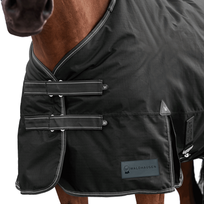 Detailaufnahme des doppelten Brustverschlusses der Waldhausen Outdoordecke Arctic 50 g in Schwarz. Die robusten T-Verschlüsse sind mit Klettabdeckungen geschützt, und können ein Hängenbleiben im Offenstall oder Paddock reduzieren. Der doppelte Brustverschluss ermöglicht einfaches Ein- und Ausdecken von Pferd oder Pony. Reflektierende Kontrastnähte in Weiß auf den Klettlaschen und dem Einfassband erhöhen die Sichtbarkeit bei Dunkelheit.