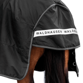 Rückansicht der dunklen Waldhausen-Outdoordecke Arctic 50 g in Schwarz mit Fokus auf den langen Schweiflatz. Der großzügige Schweiflatz schützt Schweif und Schweifrübe des Pferdes zuverlässig vor Wind, Regen und Kälte. Die breite, reflektierende Waldhausen-Applikation sorgt für optimale Sichtbarkeit von hinten im Dunkeln, auf der Weide, auf Wegen oder im Straßenverkehr. Die perfekte 600D-Ripstop-Regendecke für bessere Sichtbarkeit im Dunklen. D-Ringe für Beinschnüre sind zu sehen.