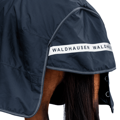 Rückansicht der dunkelblauen Waldhausen-Outdoordecke Arctic Light in Nachtblau mit Fokus auf den langen Schweiflatz. Der großzügige Schweiflatz schützt Schweif und Schweifrübe des Pferdes zuverlässig vor Wind, Regen und Kälte. Die breite, reflektierende Waldhausen-Applikation sorgt für optimale Sichtbarkeit von hinten im Dunkeln, auf der Weide, auf Wegen oder im Straßenverkehr. Die perfekte 600D-Ripstop-Regendecke für bessere Sichtbarkeit im Dunklen. D-Ringe für Beinschnüre sind zu sehen.