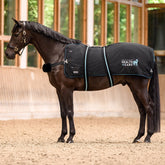Dunkelbraunes Pferd mit der schwarzen Waldhausen Health + Care TecSupreme Heiz- und Massagedecke auf dem Sandboden einer Reithalle. Die Aufnahme zeigt die Decke im praktischen Einsatz beim Warm-up vor dem Training mit sicherem Sitz über Rückenlinie, Widerrist und Kruppe.