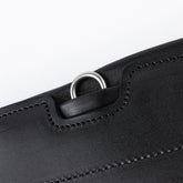Makro-Detailansicht des seitlichen Edelstahl-D-Rings am schwarzen Winderen Pressure-Relief Sattelgurt mit kleinem Bauchlatz. Der flach in das Vollnarbenleder eingearbeitete D-Ring unterhalb des Elastikzugs dient der sicheren und schnellen Befestigung von Vorderzeugen, während die versenkte Lederabdeckung ein Scheuern oder Verfangen verhindert.