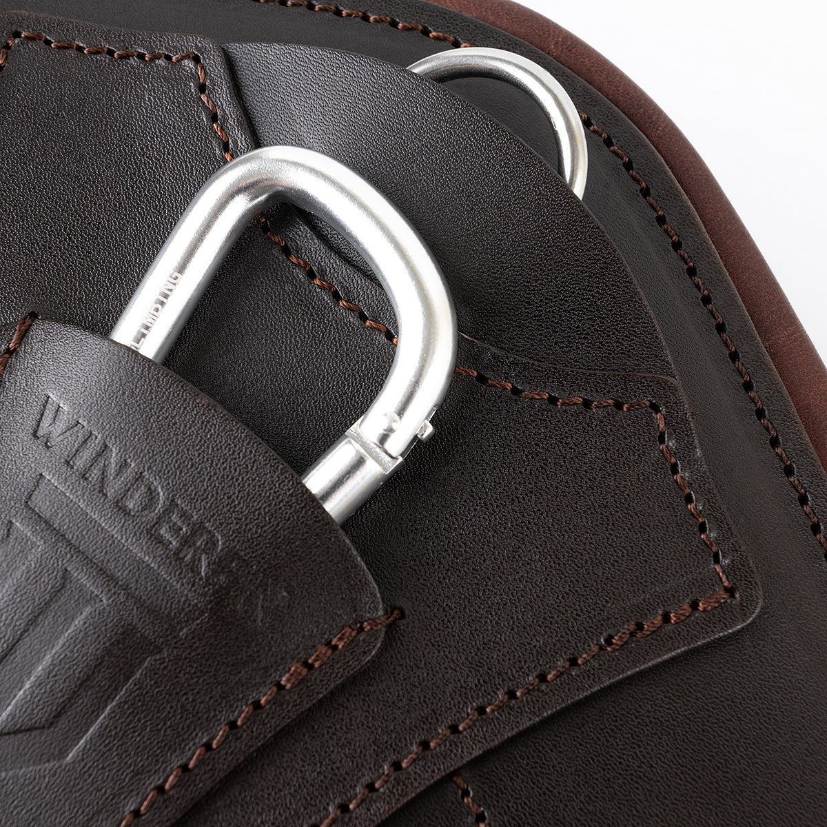 Nahaufnahme des halb versenkten Aluminium-Karabiners und D-Rings unter der Lederabdeckung mit geprägtem Winderen-Logo. Die Detailansicht zeigt die präzisen Nähte des braunen Vollnarbenleders zur hakenfreien Befestigung von Vorderzeug und Martingal.