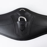 Detailansicht des schwarzen Winderen Pressure-Relief Springgurtes mit kleinem Bauchlatz aus hochwertigem Vollnarbenleder. Die Nahaufnahme zeigt die Leder-Schutzabdeckung mit geprägtem Winderen-Logo über dem halb versenkten Aluminium-Karabiner für das Einhaken von Vorderzeugen sowie den darüber liegenden Edelstahl-D-Ring für Hilfszügel wie Martingale.