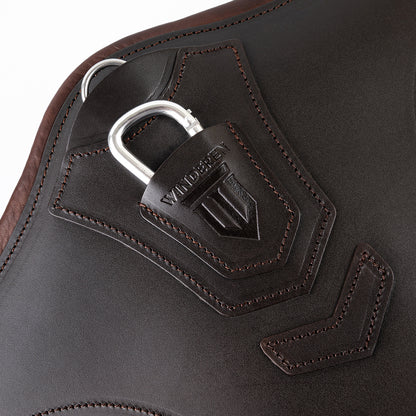 Detailaufnahme des braunen Winderen-Pressure-Relief-Stollengurts aus Vollnarbenleder mit eingeprägtem Winderen-Logo. Zu sehen sind der halb versenkte Aluminium-Karabiner unter einer Schutzabdeckung aus Leder zum sicheren Einhaken des Vorderzeugs sowie der darüber liegende D-Ring für Hilfszügel wie das Martingal. Die präzisen Nähte und die flache Lederverarbeitung verhindern das Verfangen über dem Sprung. Perfekt durchdachtes Premium-Sattelzubehör für das Springreiten.